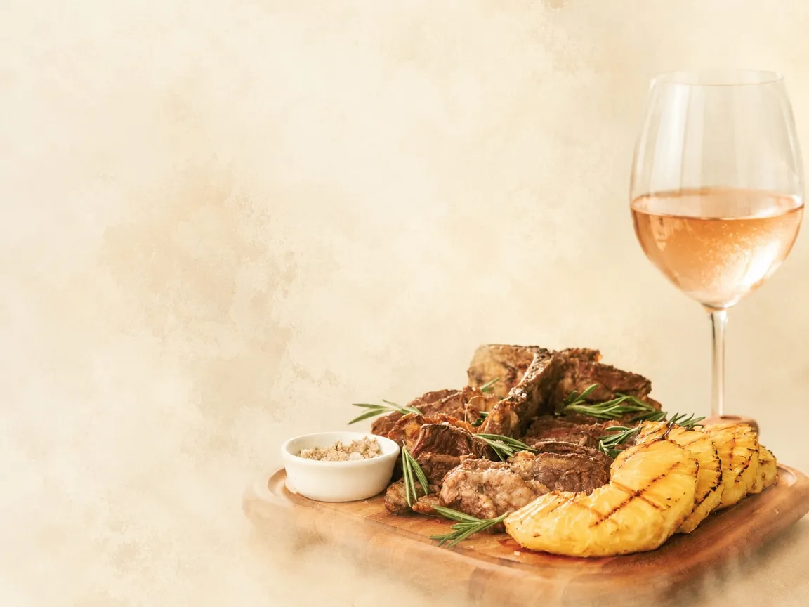 Tábua de carne grelhada com ananás assado, alecrim e flor de sal, e um copo de vinho rosé, sobre fundo creme